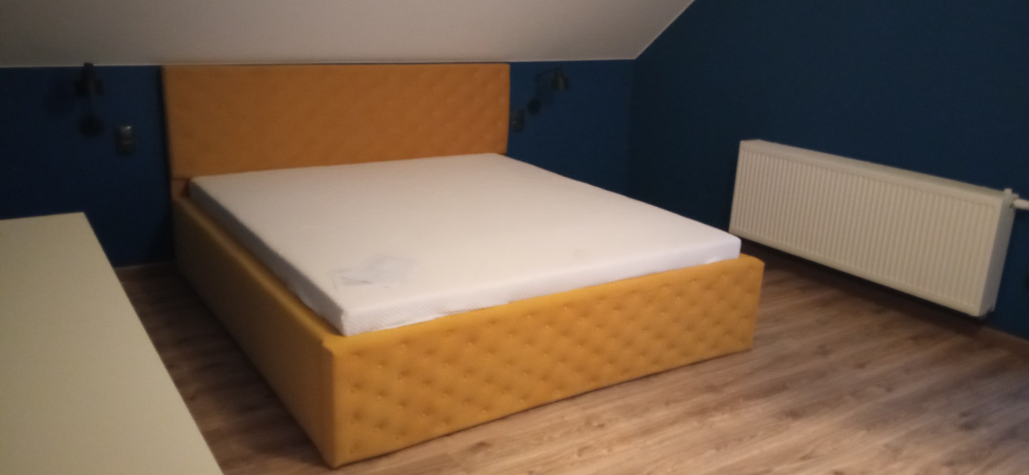 Żółte łóżko tapicerowane z pikowanym wezgłowiem i ramą, na nim biały materac, w pokoju z granatową ścianą i białym grzejnikiem.