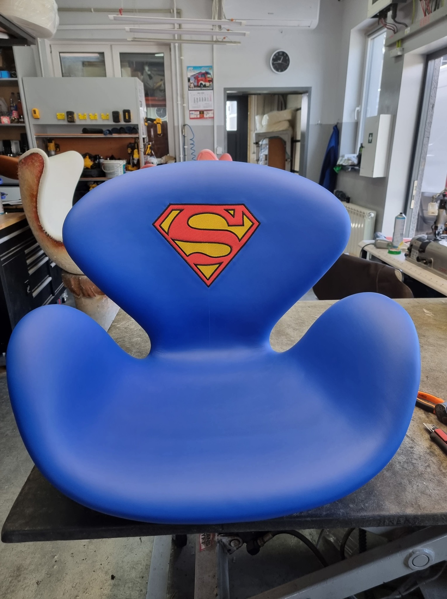 Niebieski fotel dziecięcy w kształcie litery S z logo Supermana, stojący na stole w warsztacie tapicerskim, widoczne narzędzia i maszyna do szycia w tle.