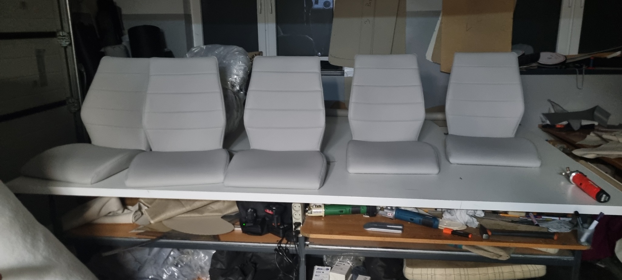 Cztery białe, tapicerowane siedzenia samochodowe z oparciami, ułożone obok siebie na białym stole warsztatowym, widoczne narzędzia i materiały tapicerskie w tle.