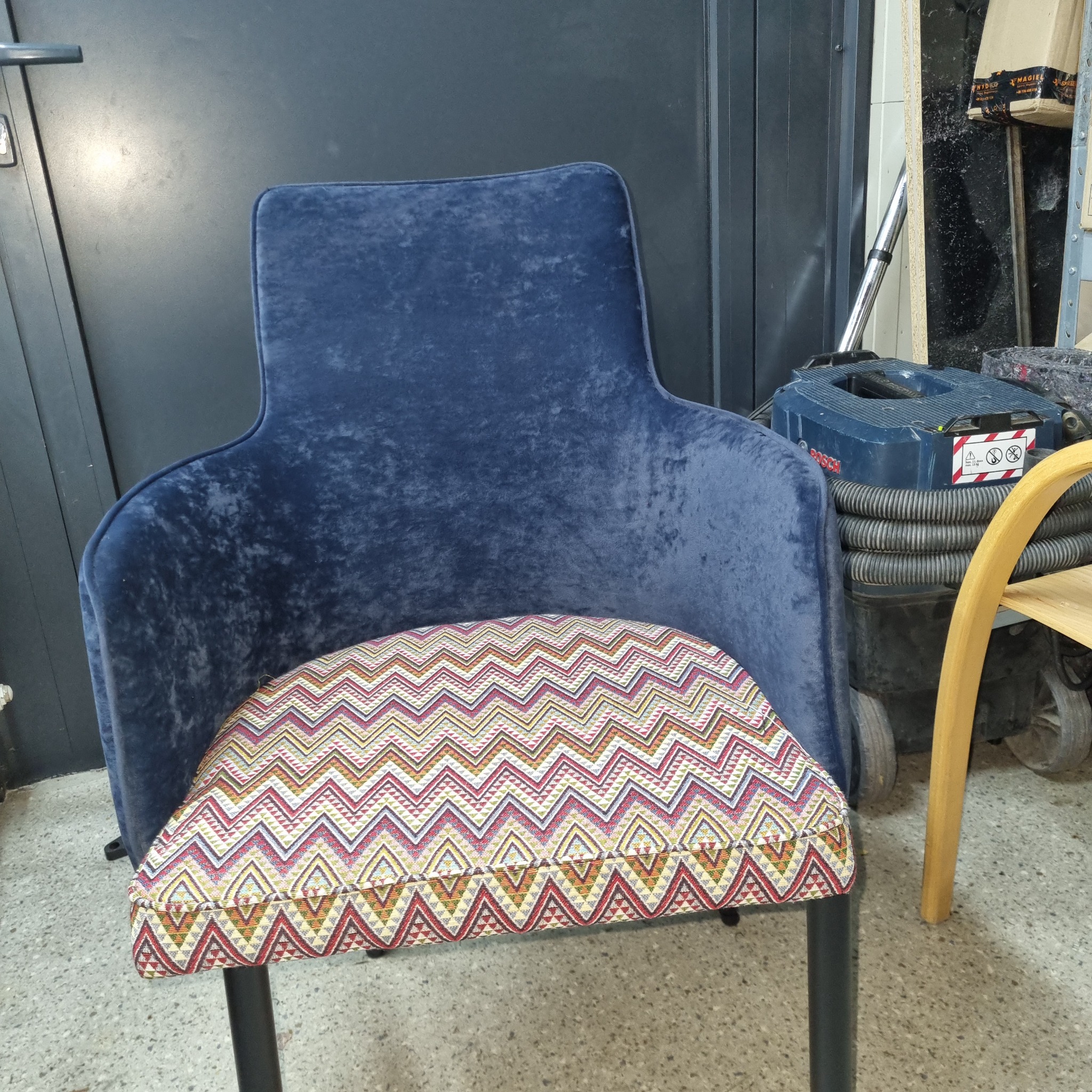 Krzesło z ciemnoniebieskim, welurowym obiciem i wzorzystym siedziskiem w zygzaki, stojące na tle szarej ściany i sprzętu warsztatowego.