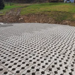 Ułożona szara ażurowa nawierzchnia z betonowych płyt z okrągłymi otworami, częściowo podsypana szarym żwirem, na tle ziemnego nasypu porośniętego trawą i drzewami.