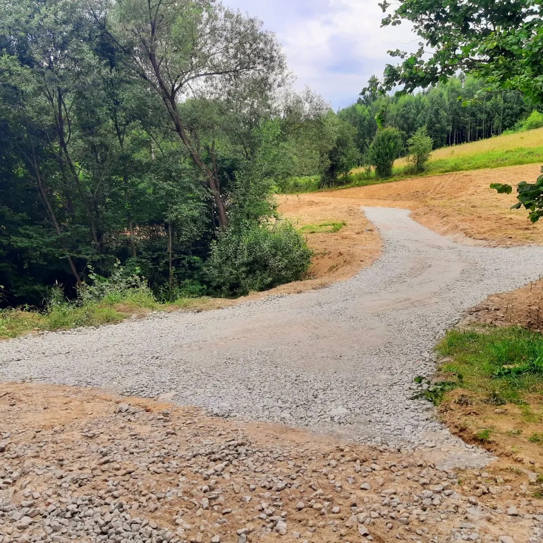 Nowo utwardzona, kręta droga żwirowa prowadząca przez teren przygotowany pod budowę, otoczona zielenią i drzewami.