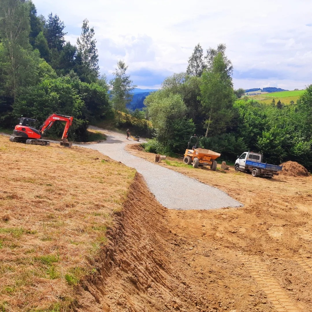 Wyrównany teren pod budowę drogi gruntowej na zboczu, widoczny czerwony mini-koparka, mała wywrotka i ciężarówka, w tle zalesione wzgórza.