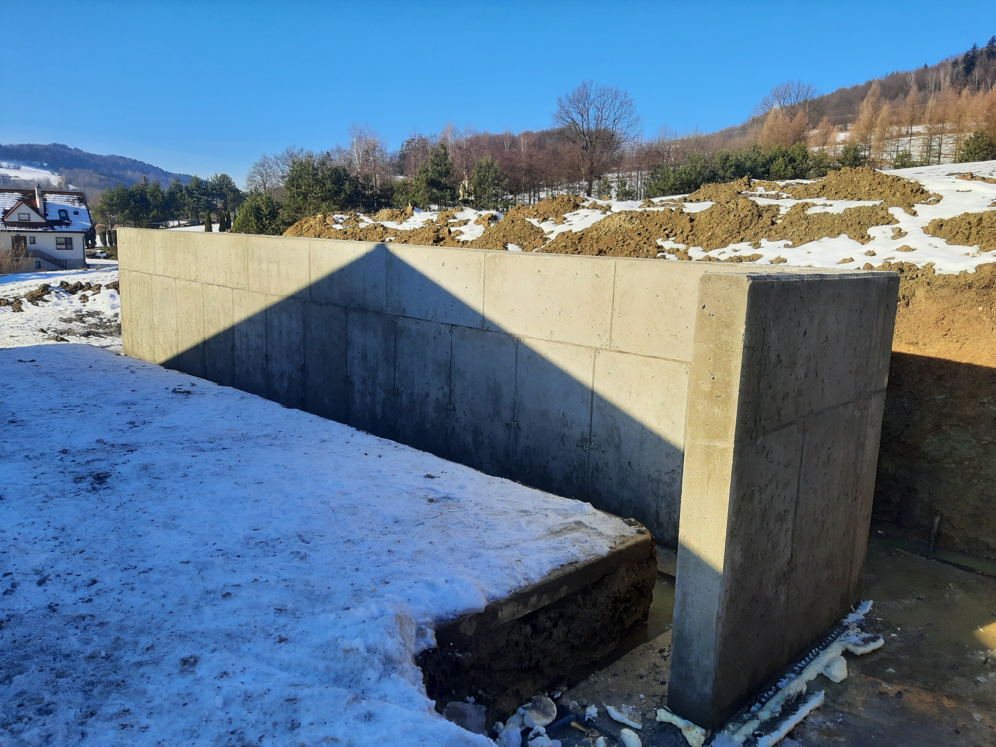 Betonowy fundament w zimowej scenerii, częściowo przysypany śniegiem, z widocznym wykopem i ziemią w tle, na tle zalesionych wzgórz pod błękitnym niebem.