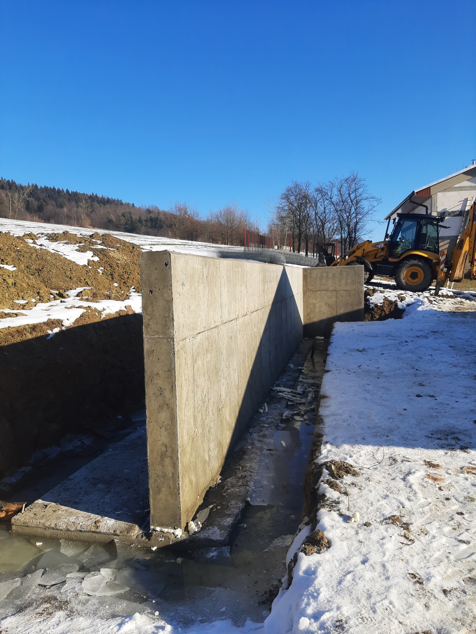Wzmocniony, betonowy fundament ściany oporowej w wykopie, z częściowo zamarzniętą wodą i śniegiem, na tle krajobrazu zimowego i żółtej koparko-ładowarki.