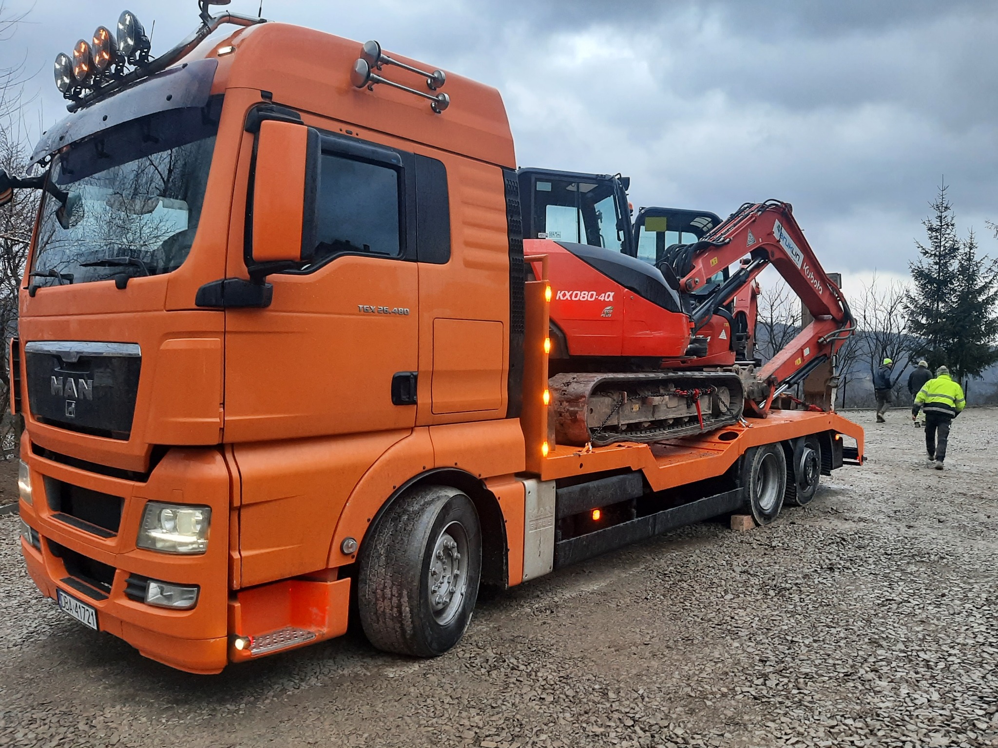 Pomarańczowy transportowiec MAN z niskopodwoziową naczepą przewozi dwie czerwone mini-koparki Kubota KX080-4α na żwirowym podłożu, widoczni pracownicy w odblaskowych kamizelkach.