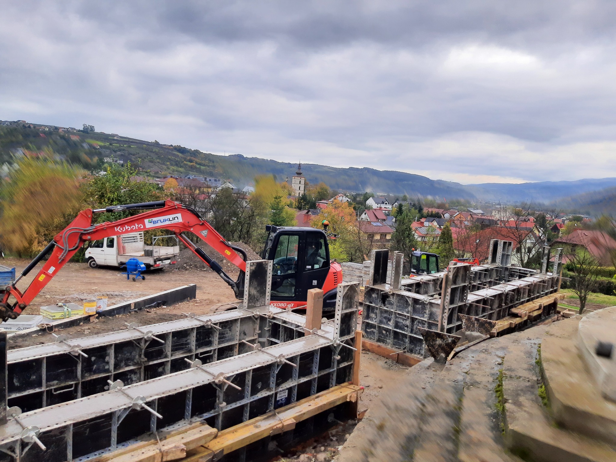 Szare metalowe szalunki przygotowane do zalewania betonem, czerwona koparka Kubota z przodu, w tle zabudowa miejska z wieżą kościelną i pagórkowaty krajobraz pod pochmurnym niebem.