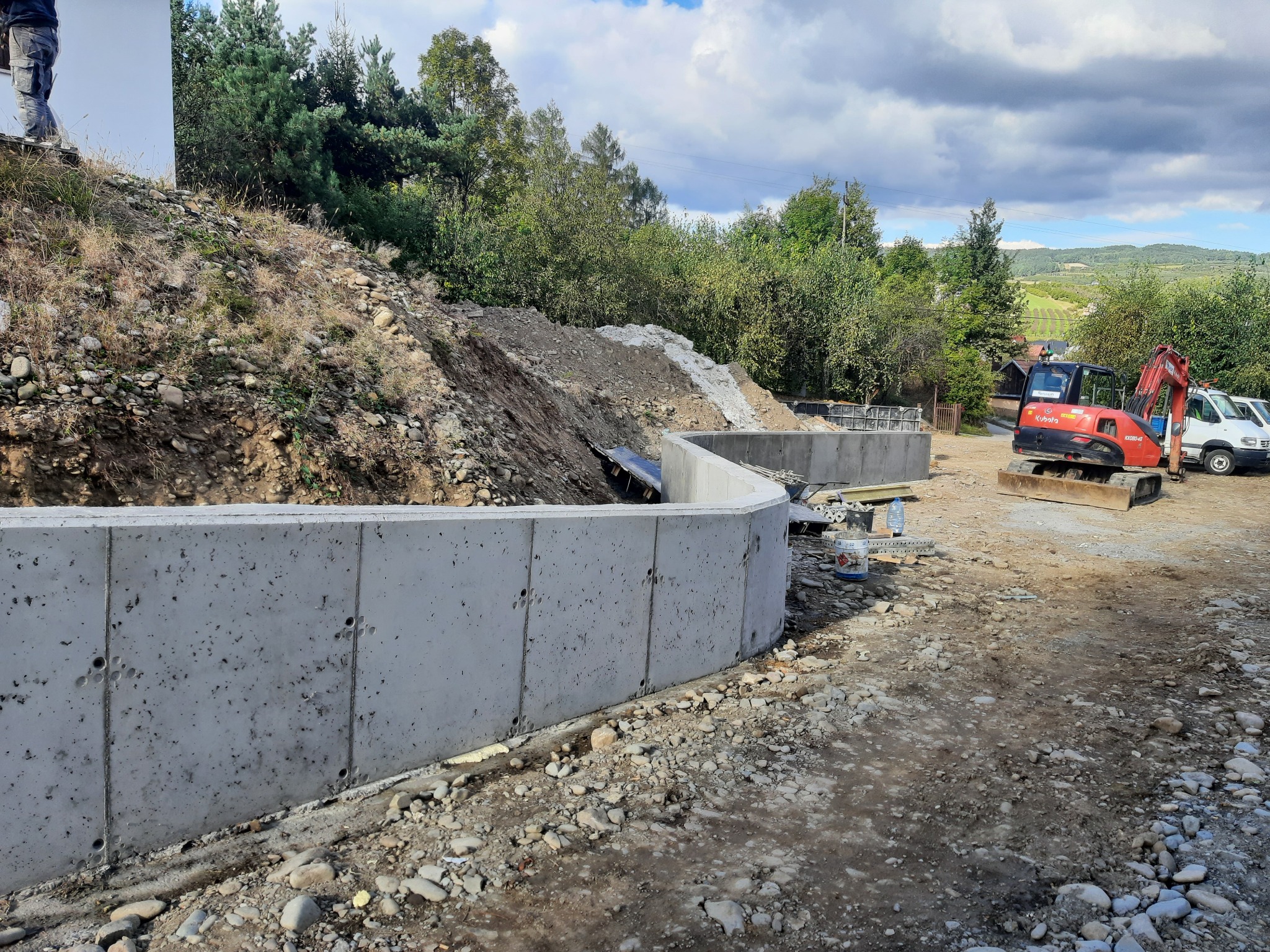 Betonowy mur oporowy w trakcie budowy, widoczny wykop ziemny, mała koparka Kubota i samochód dostawczy w tle, teren budowy z kamieniami i ziemią.