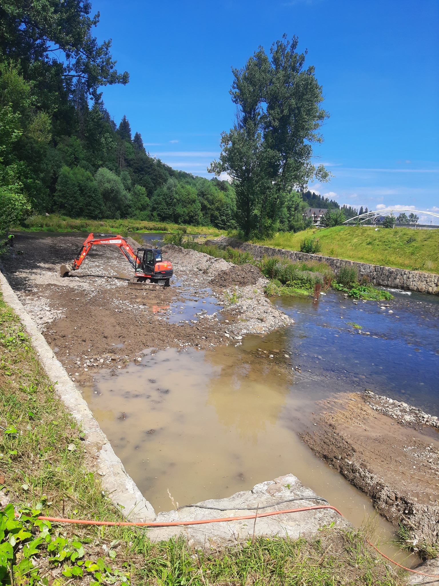 Pomarańczowa koparka Kubota na błotnistym brzegu rzeki, w tle zieleń i błękitne niebo, prace ziemne w korycie rzeki