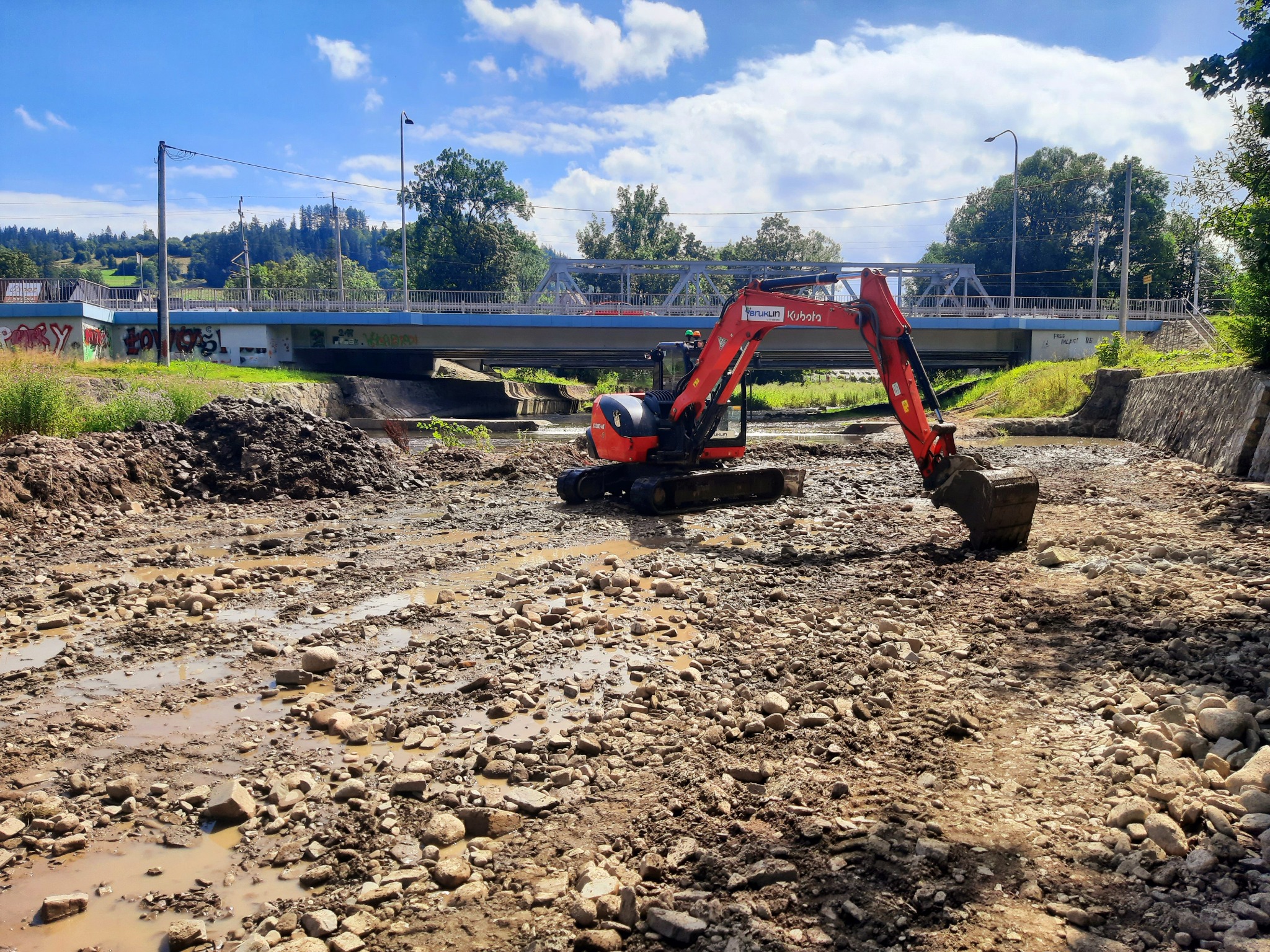 Czerwona koparka Kubota w korycie rzeki z kamienistym dnem, pracująca przy regulacji rzeki pod stalowym mostem z graffiti.