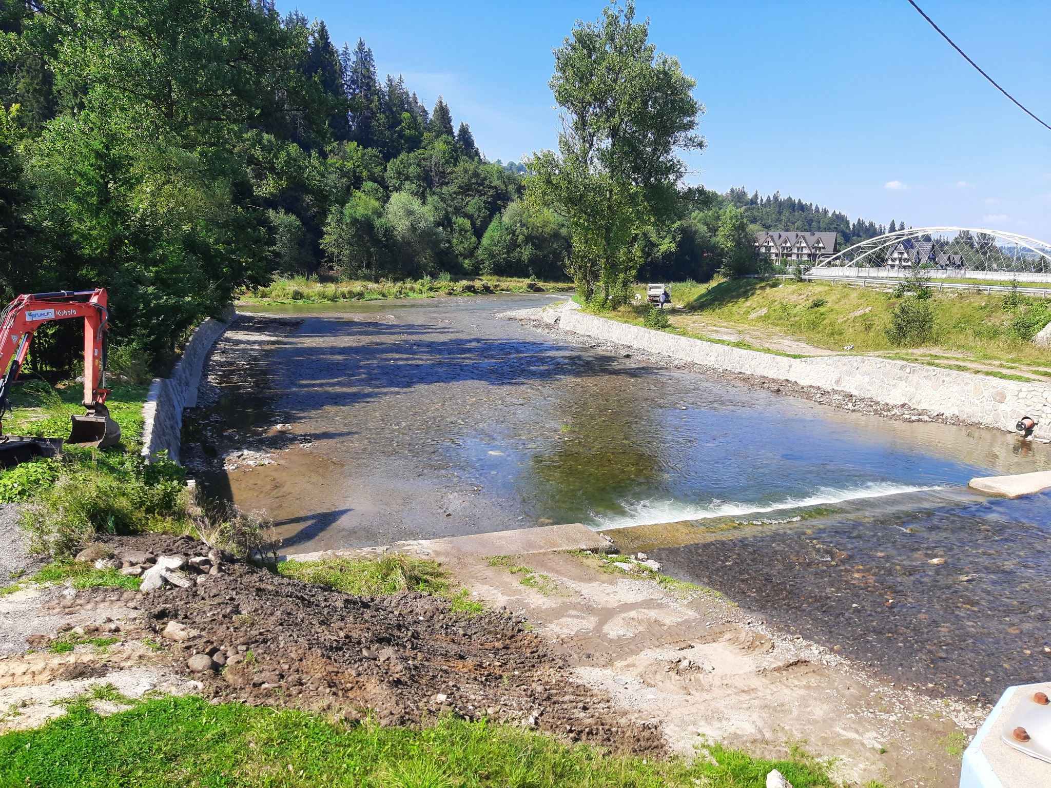 Regulacja rzeki z użyciem betonowych umocnień i małej kaskady, widoczny mały czerwony koparka na brzegu, w tle zalesione wzgórza i zabudowania.