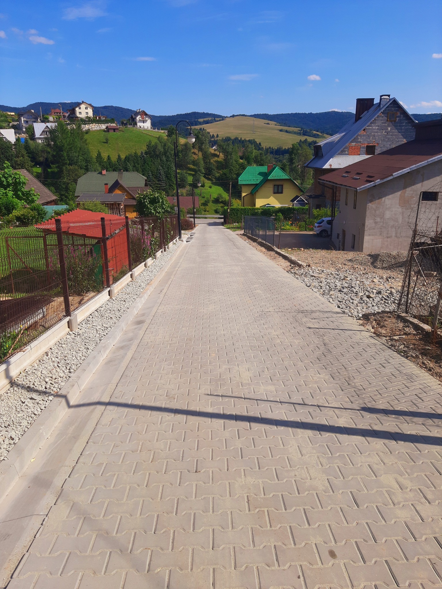 Nowa, wybrukowana droga z szarej kostki betonowej w górskiej scenerii, z widocznymi domami i zielonymi wzgórzami w tle, oświetlona silnym słońcem rzucającym cienie słupów.