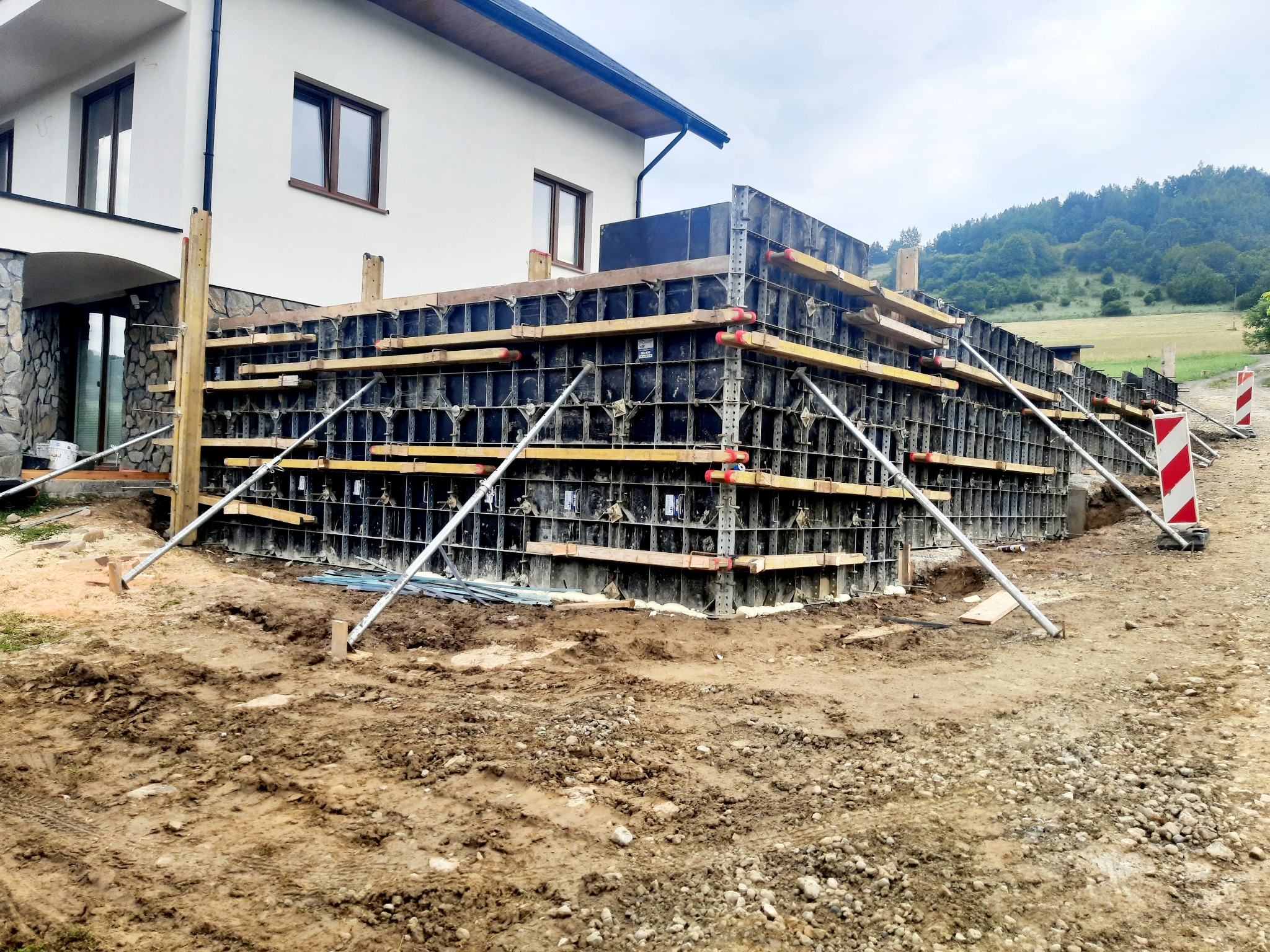 Szare szalunki fundamentowe z drewnianymi belkami i metalowymi wspornikami na tle budowanego domu jednorodzinnego z kamienną elewacją i zielonym krajobrazem w tle.