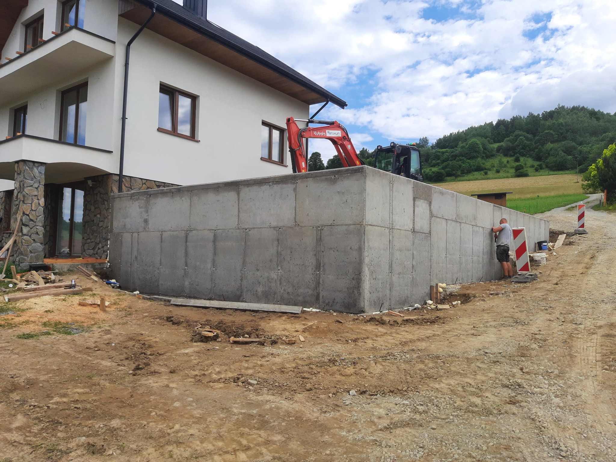 Wylany betonowy fundament domu jednorodzinnego z widocznym minikoparką Kubota i pracownikiem przy ścianie, na tle pagórkowatego krajobrazu.