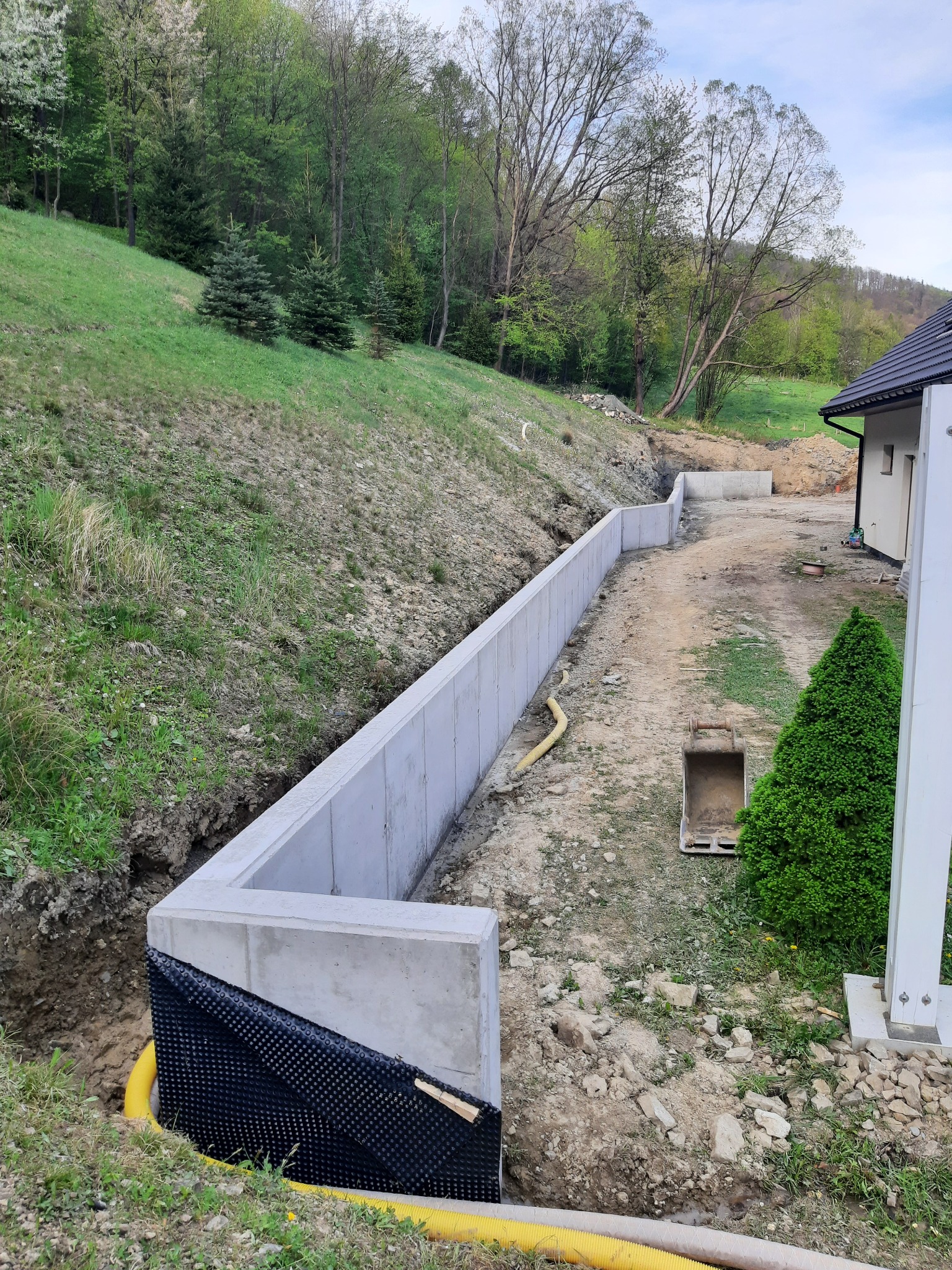 Wzniesiony betonowy fundament z izolacją pionową z folii kubełkowej i rurą drenażową, widok na wykop i zbocze porośnięte trawą oraz fragment domu w tle.