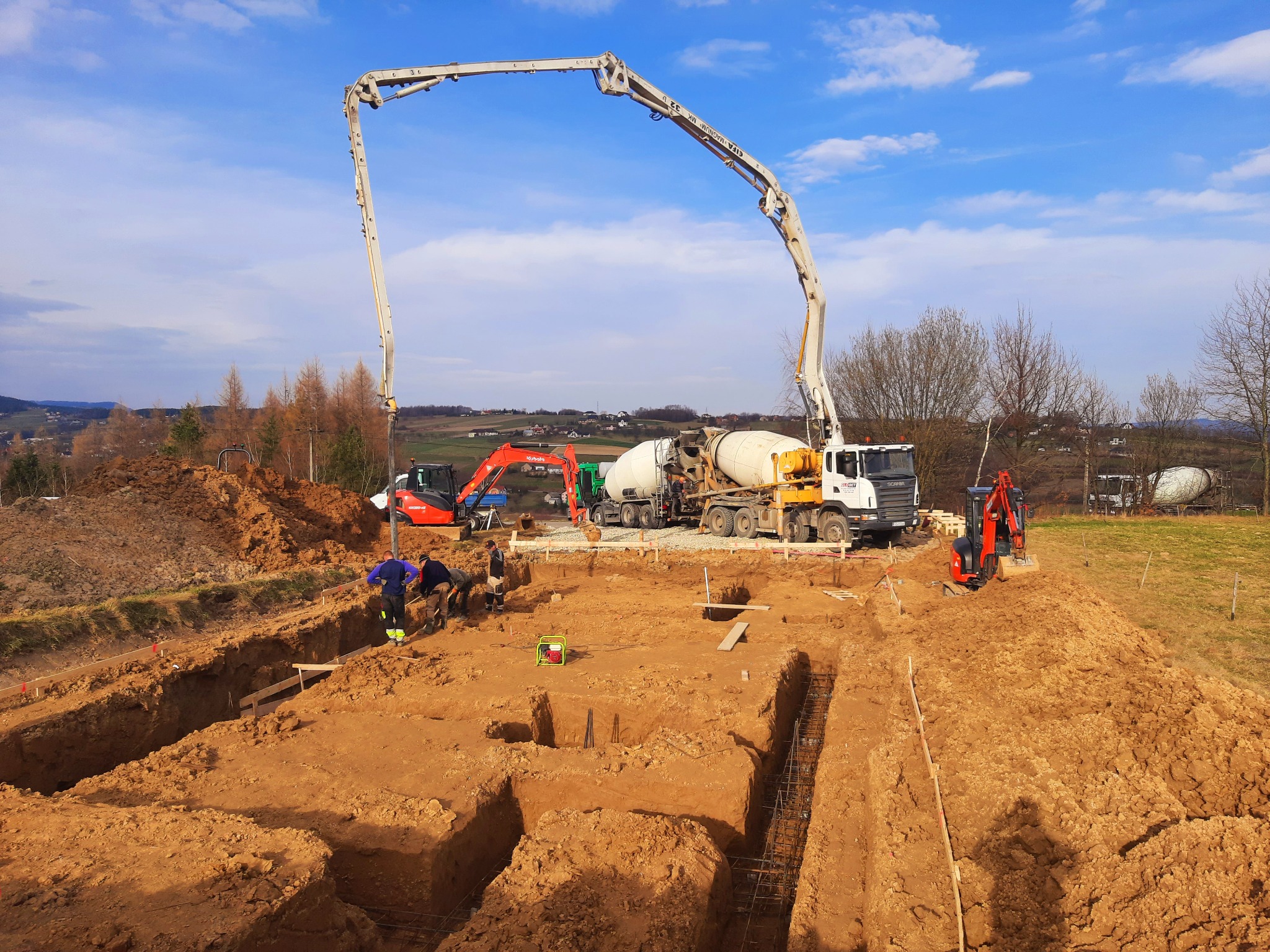 Wykopane rowy pod fundamenty z ułożonym zbrojeniem, widoczni pracownicy, pompa do betonu oraz betoniarka na placu budowy w słoneczny dzień.
