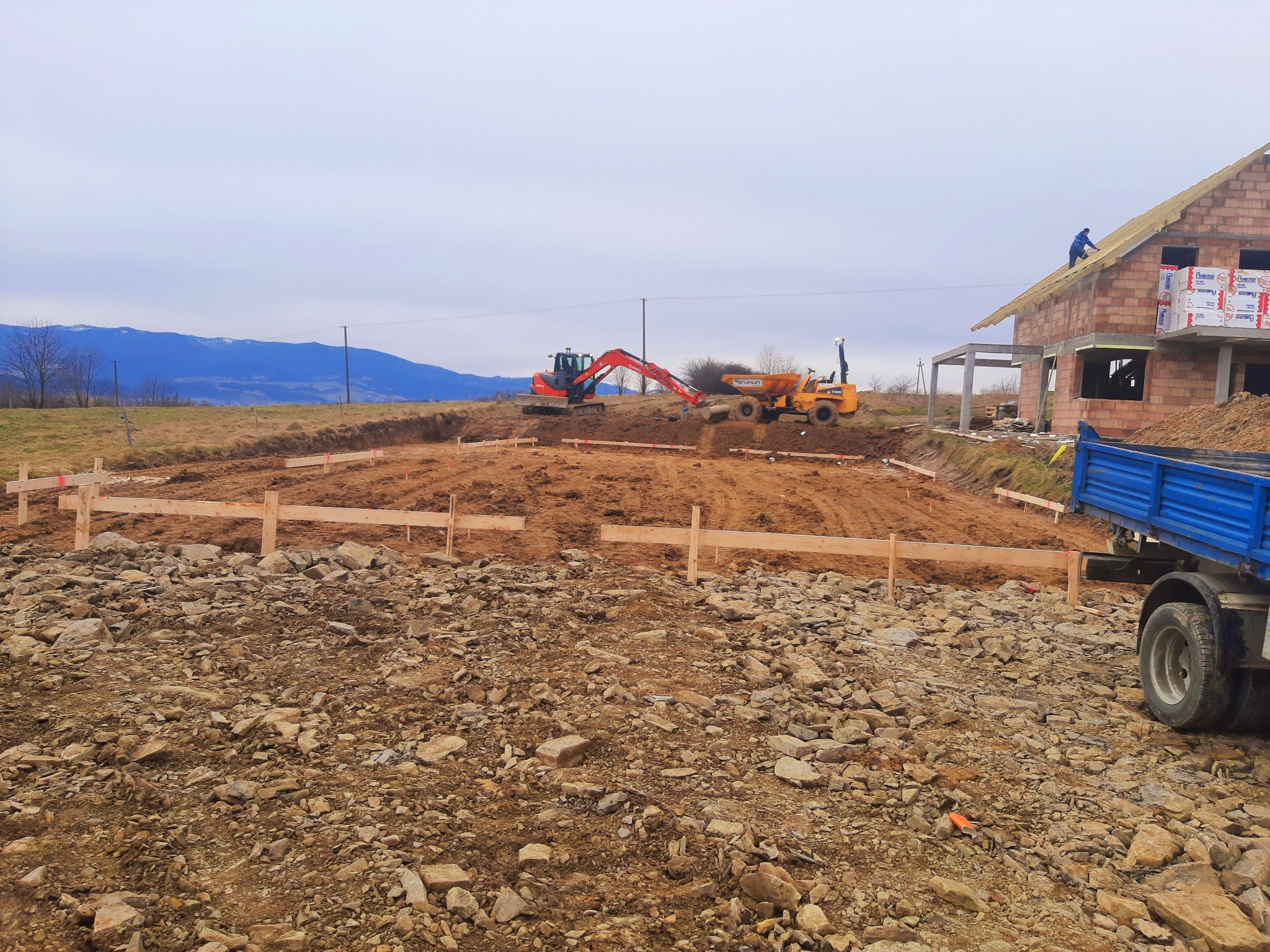Przygotowanie terenu pod budowę domu: wykopane fundamenty z drewnianym szalunkiem, pracujący koparka i wywrotka, w tle budynek w trakcie budowy z czerwonej cegły i ociepleniem.