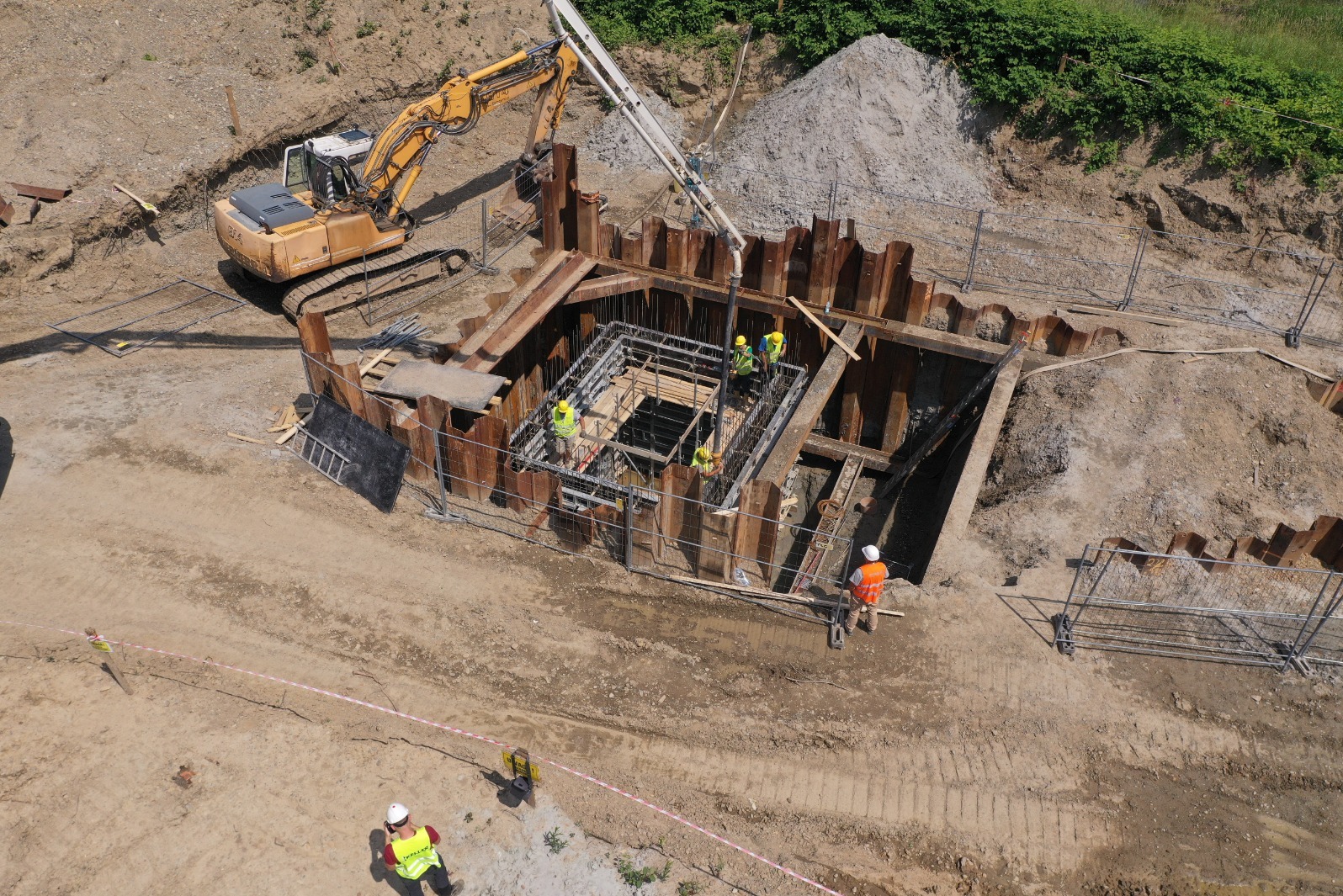 Z góry widok na budowę zabezpieczonego wykopu z pracującymi robotnikami w żółtych kamizelkach, pompą do betonu i koparką na placu budowy.
