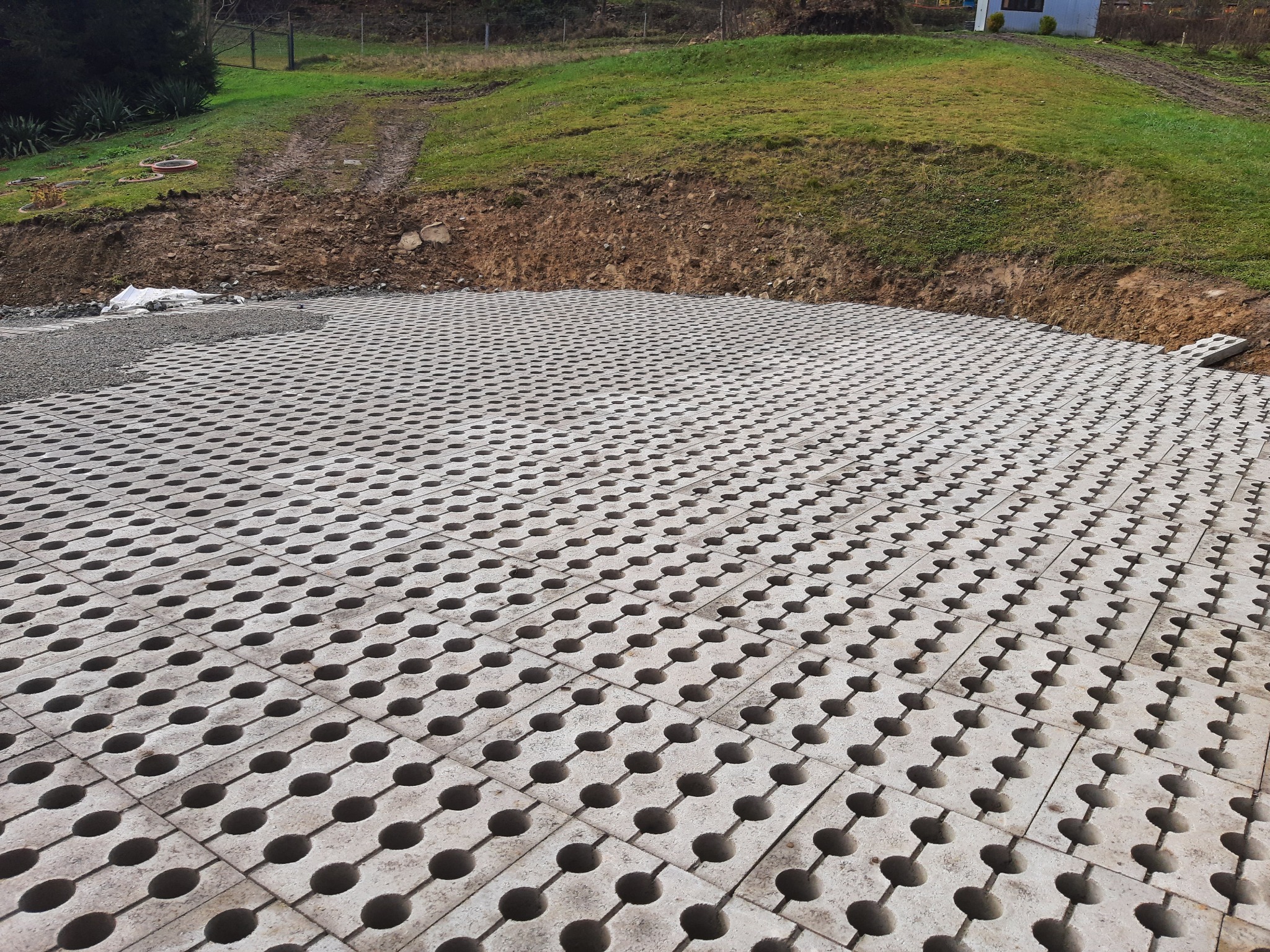 Ułożona szara ażurowa nawierzchnia z betonowych płyt z okrągłymi otworami, częściowo podsypana szarym żwirem, na tle ziemnego nasypu porośniętego trawą i drzewami.