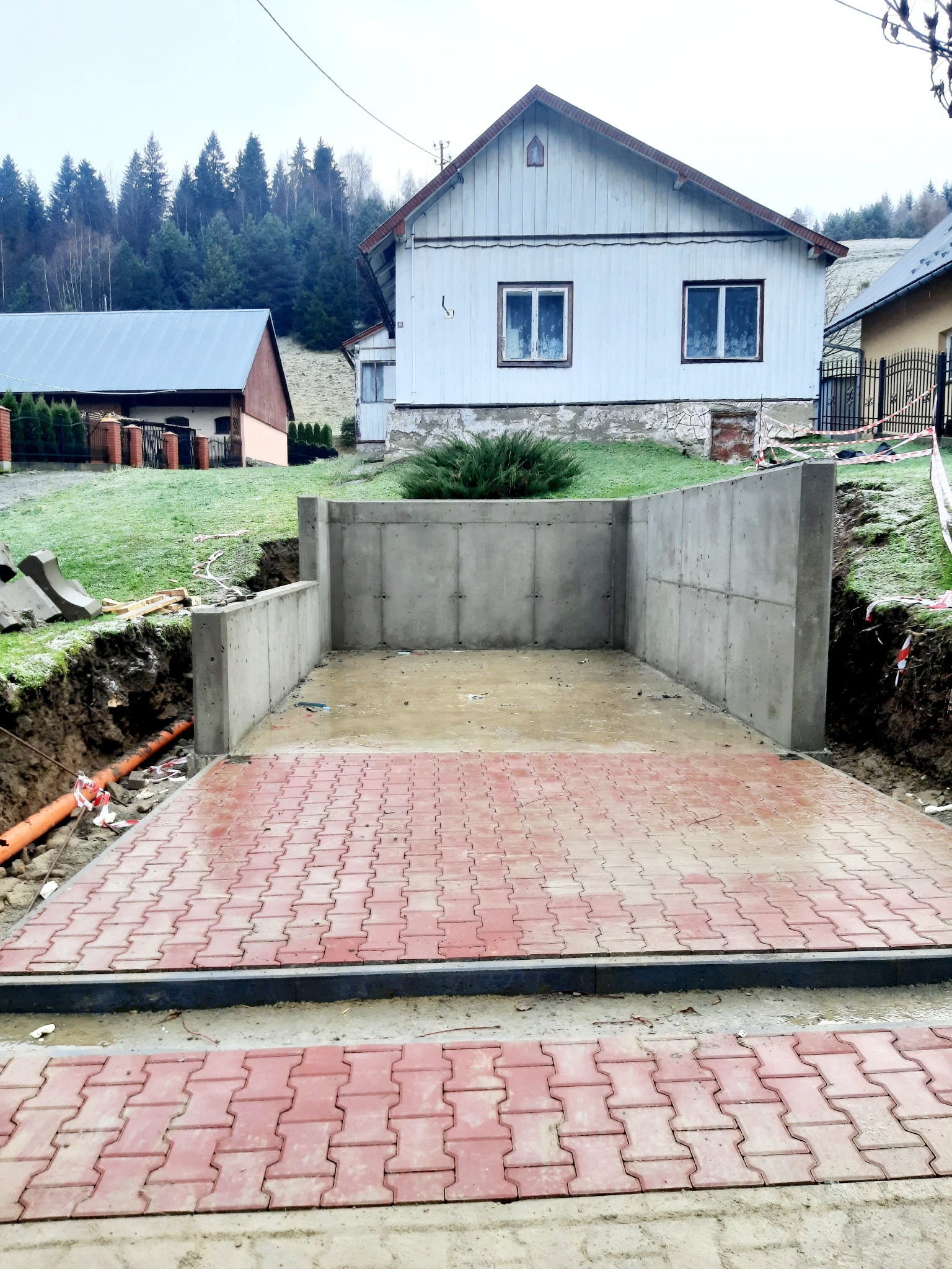 Betonowa konstrukcja w ziemi z ułożoną czerwoną kostką brukową, widoczne rury i wykop, w tle domy i zalesione wzgórze.