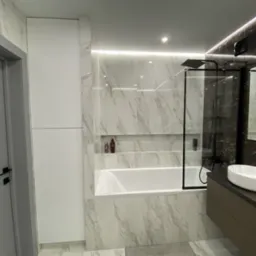 Nowoczesna łazienka z białą wanną, marmurowymi płytkami na ścianach, kabiną prysznicową z czarnymi ramami i szafką z umywalką w kolorze taupe.