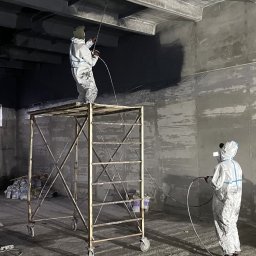 ZŁOTY KĄT SPÓŁKA Z OGRANICZONĄ ODPOWIEDZIALNOŚCIĄ - Dwóch pracowników w kombinezonach ochronnych maluje natryskowo sufit i ściany dużego pomieszczenia, jeden stoi na rusztowaniu, na podłodze leży drabina.