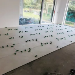 Jasne płytki podłogowe w trakcie układania, z użyciem systemu poziomowania płytek (zielone klipsy i kliny), widoczne duże okna i surowy beton na podłodze w tle.