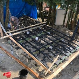 ZŁOTY KĄT SPÓŁKA Z OGRANICZONĄ ODPOWIEDZIALNOŚCIĄ - Budowa tarasu: czarna, pomalowana konstrukcja z belek na zielonej izolacji, widoczny mężczyzna w kraciastej koszuli przy pracy, drzewa w tle.