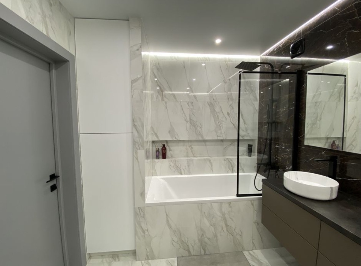 Nowoczesna łazienka z białą wanną, marmurowymi płytkami na ścianach, kabiną prysznicową z czarnymi ramami i szafką z umywalką w kolorze taupe.