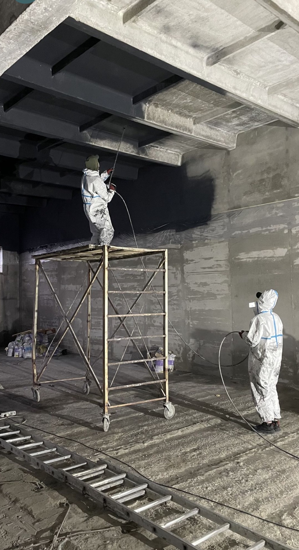 Dwóch pracowników w kombinezonach ochronnych maluje natryskowo sufit i ściany dużego pomieszczenia, jeden stoi na rusztowaniu, na podłodze leży drabina.