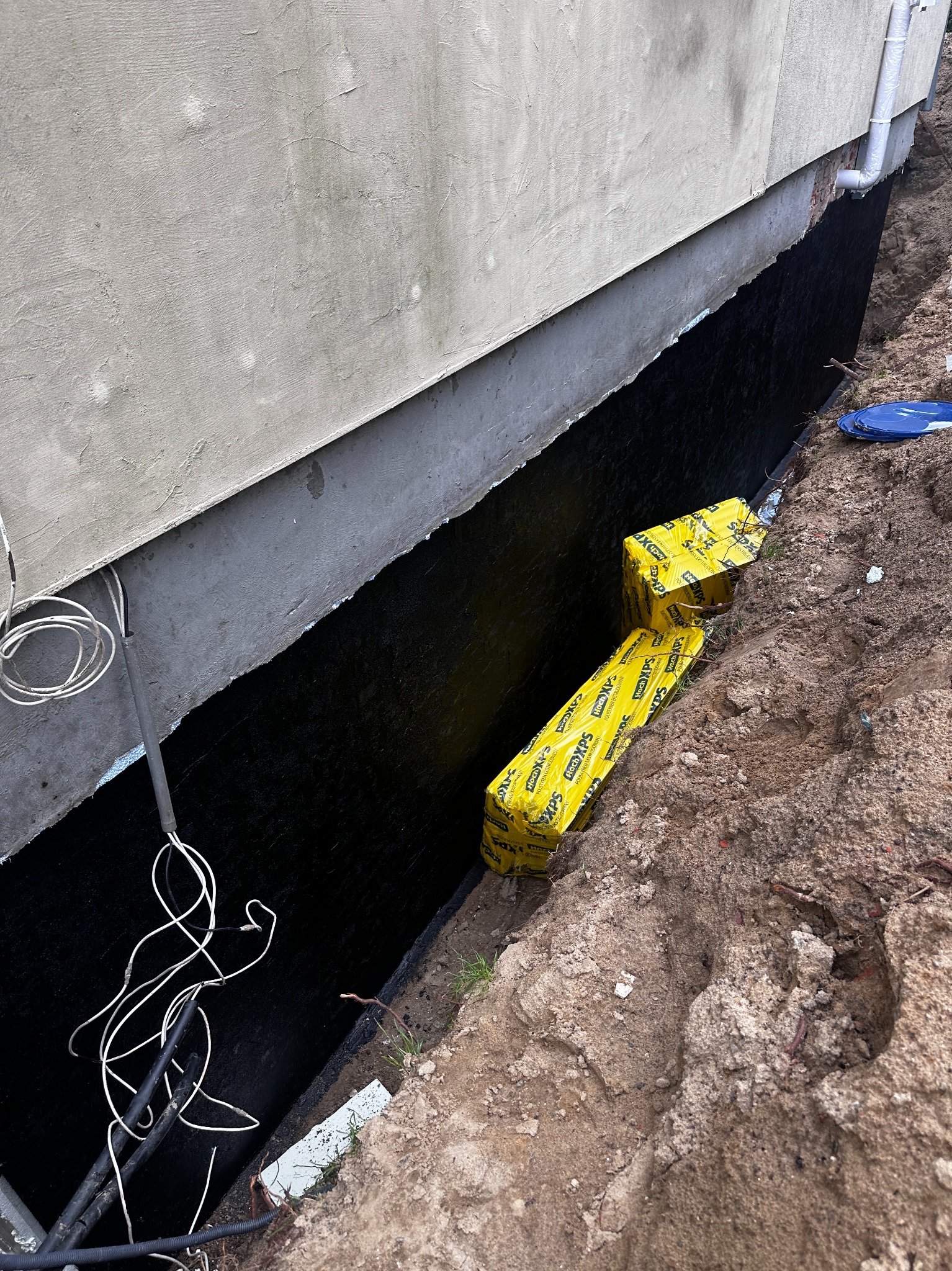 Izolacja fundamentów budynku w trakcie prac: czarna powłoka hydroizolacyjna, żółte opakowania styropianu XPS Hoch XPS, kable elektryczne przy ścianie, wykop ziemny.