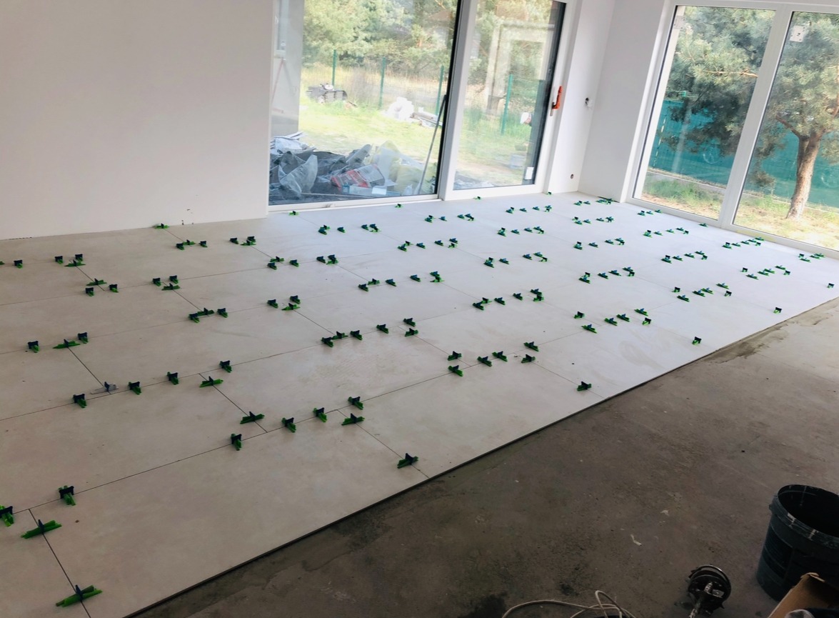 Jasne płytki podłogowe w trakcie układania, z użyciem systemu poziomowania płytek (zielone klipsy i kliny), widoczne duże okna i surowy beton na podłodze w tle.