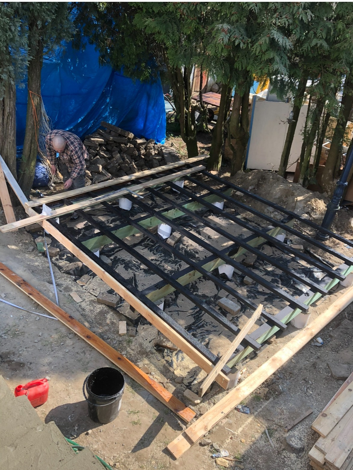 Budowa tarasu: czarna, pomalowana konstrukcja z belek na zielonej izolacji, widoczny mężczyzna w kraciastej koszuli przy pracy, drzewa w tle.