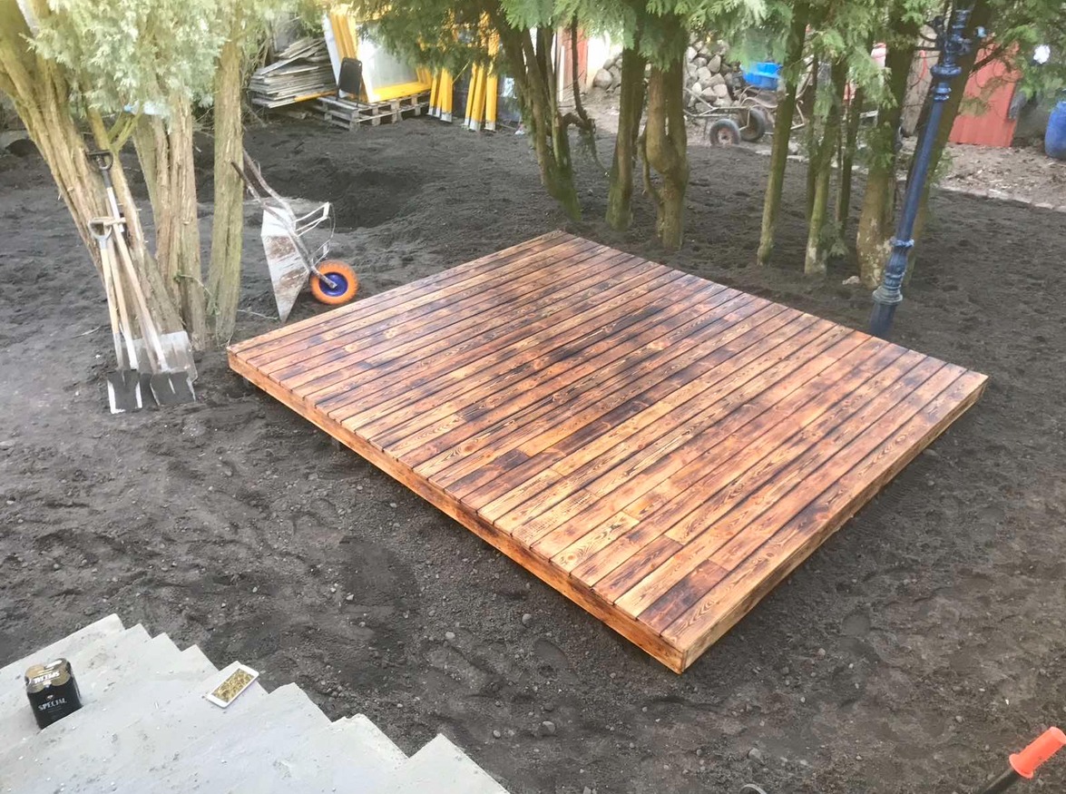 Drewniany podest ułożony na ziemi, widoczne narzędzia ogrodnicze, taczka i schody betonowe.