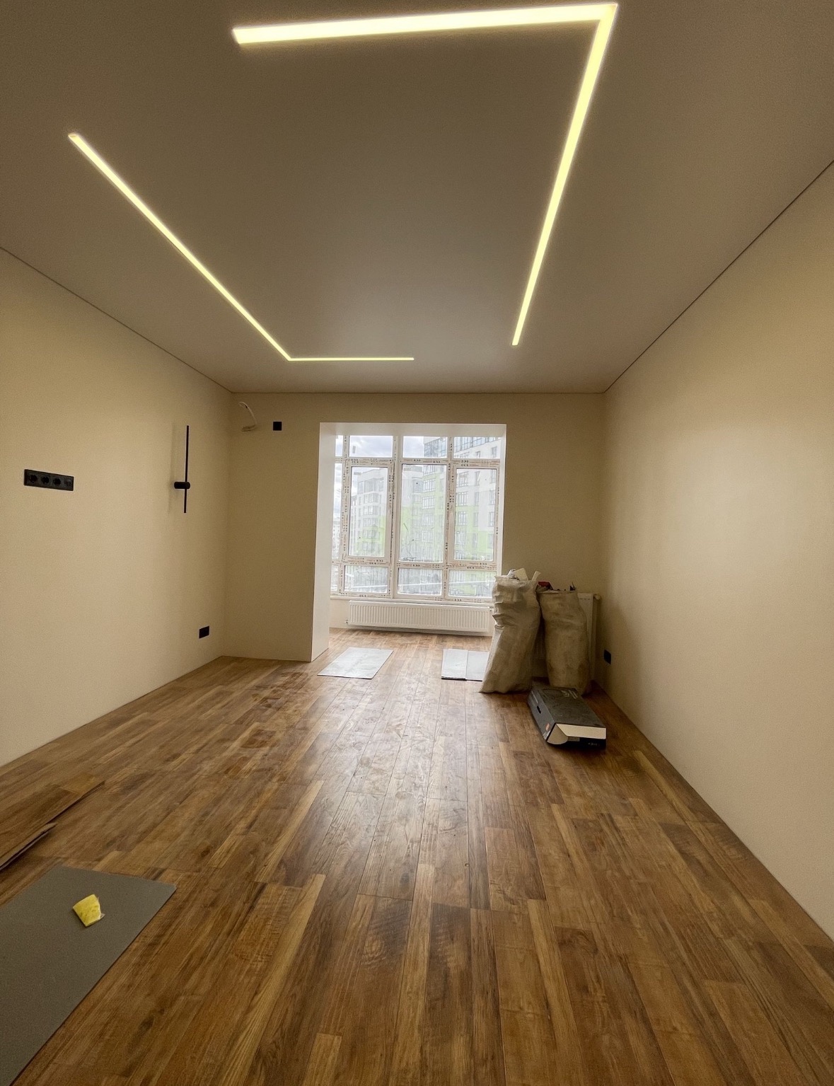 Pokój w trakcie wykończenia z drewnopodobną podłogą, workami z materiałami budowlanymi przy oknie i nowoczesnym, liniowym oświetleniem LED na suficie.