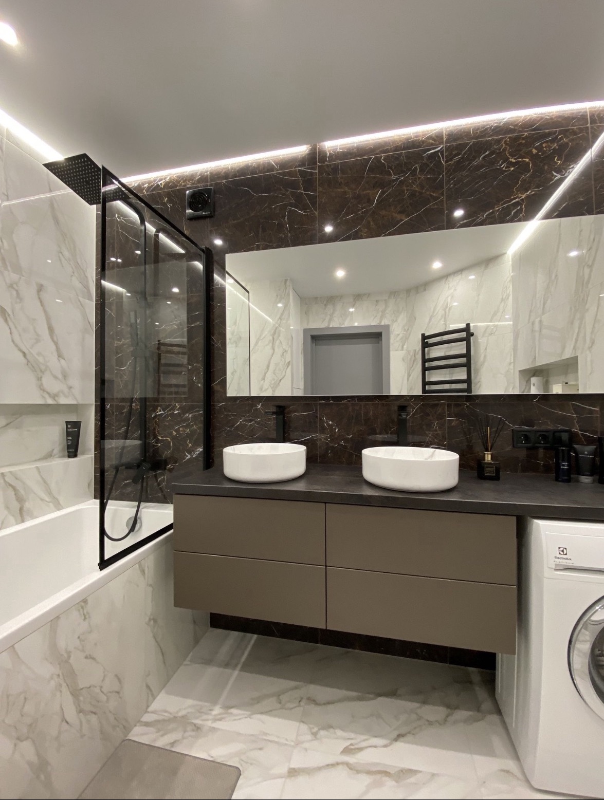 Nowoczesna łazienka z marmurowymi płytkami, dwoma białymi umywalkami nablatowymi, czarną armaturą i kabiną prysznicową typu walk-in z czarnymi ramami, oświetleniem LED i pralką Electrolux z boku.