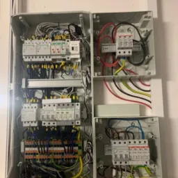 Otwarta szafa elektryczna z widocznymi zabezpieczeniami przepięciowymi, bezpiecznikami i okablowaniem w kolorach: czerwonym, żółto-zielonym, czarnym i niebieskim. Na dole widoczne etykiety...