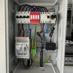 PRO TECH Tomasz Palkowski - Wewnętrzny widok skrzynki elektrycznej z zainstalowanymi bezpiecznikami, ogranicznikiem przepięć i routerem w Mysłowicach, z widocznymi przewodami w różnych kolorach.