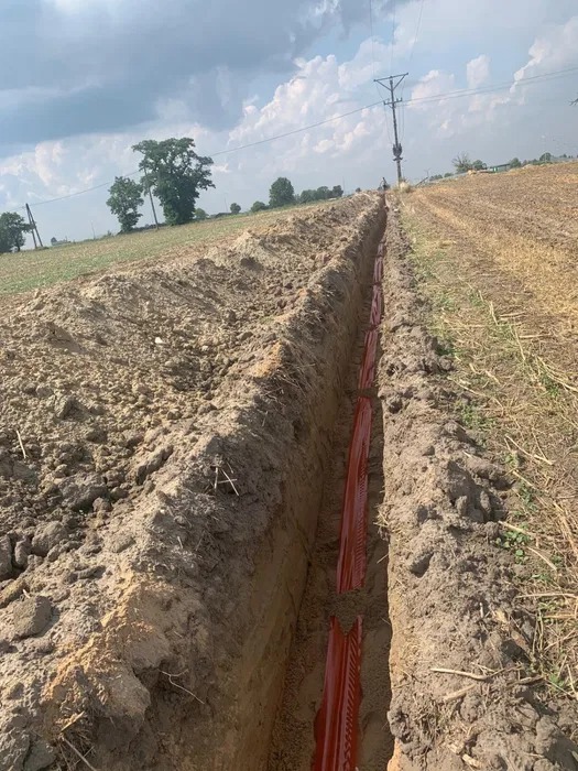 Wykop w polu z ułożonymi rurami osłonowymi dla kabli elektrycznych, widoczna linia wysokiego napięcia w oddali.