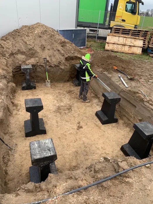 Przygotowanie fundamentów pod instalację odgromową: pracownik w odblaskowej kurtce poziomuje betonowe słupki w wykopie, widoczne narzędzia i sprzęt budowlany, w tle budynek przemysłowy i ciężarówka.