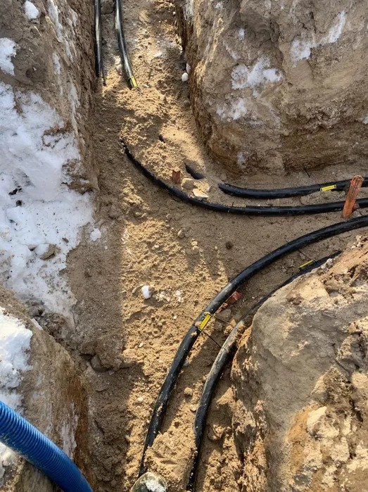 Wykop z ułożonymi przewodami elektrycznymi w ziemi, widoczne oznaczenia na kablach i resztki śniegu na ścianach wykopu.
