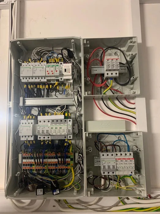 Otwarta szafa elektryczna z widocznymi zabezpieczeniami przepięciowymi, bezpiecznikami i okablowaniem w kolorach: czerwonym, żółto-zielonym, czarnym i niebieskim. Na dole widoczne etykiety...