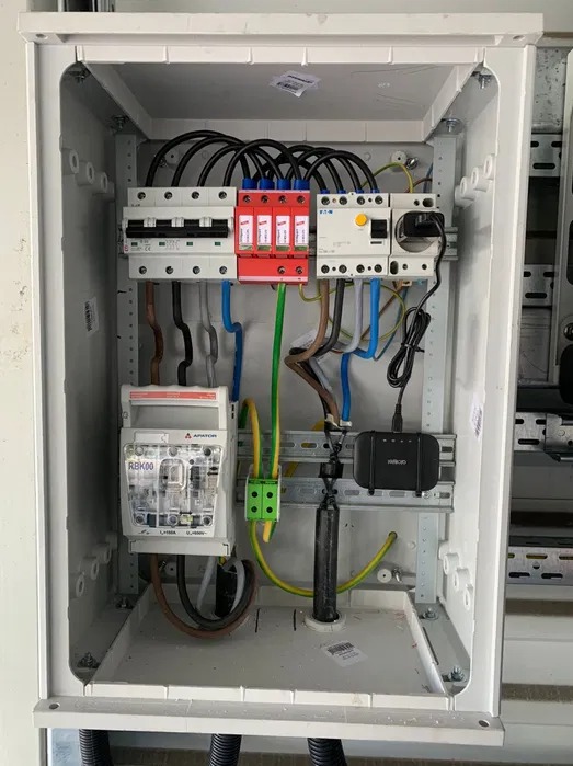 Wewnętrzny widok skrzynki elektrycznej z zainstalowanymi bezpiecznikami, ogranicznikiem przepięć i routerem w Mysłowicach, z widocznymi przewodami w różnych kolorach.