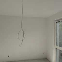 Szary kabel zwisa z sufitu w jasnym pokoju z oknem. Widoczne puszki elektryczne na ścianie. Przygotowanie do instalacji oświetlenia w nowym mieszkaniu.