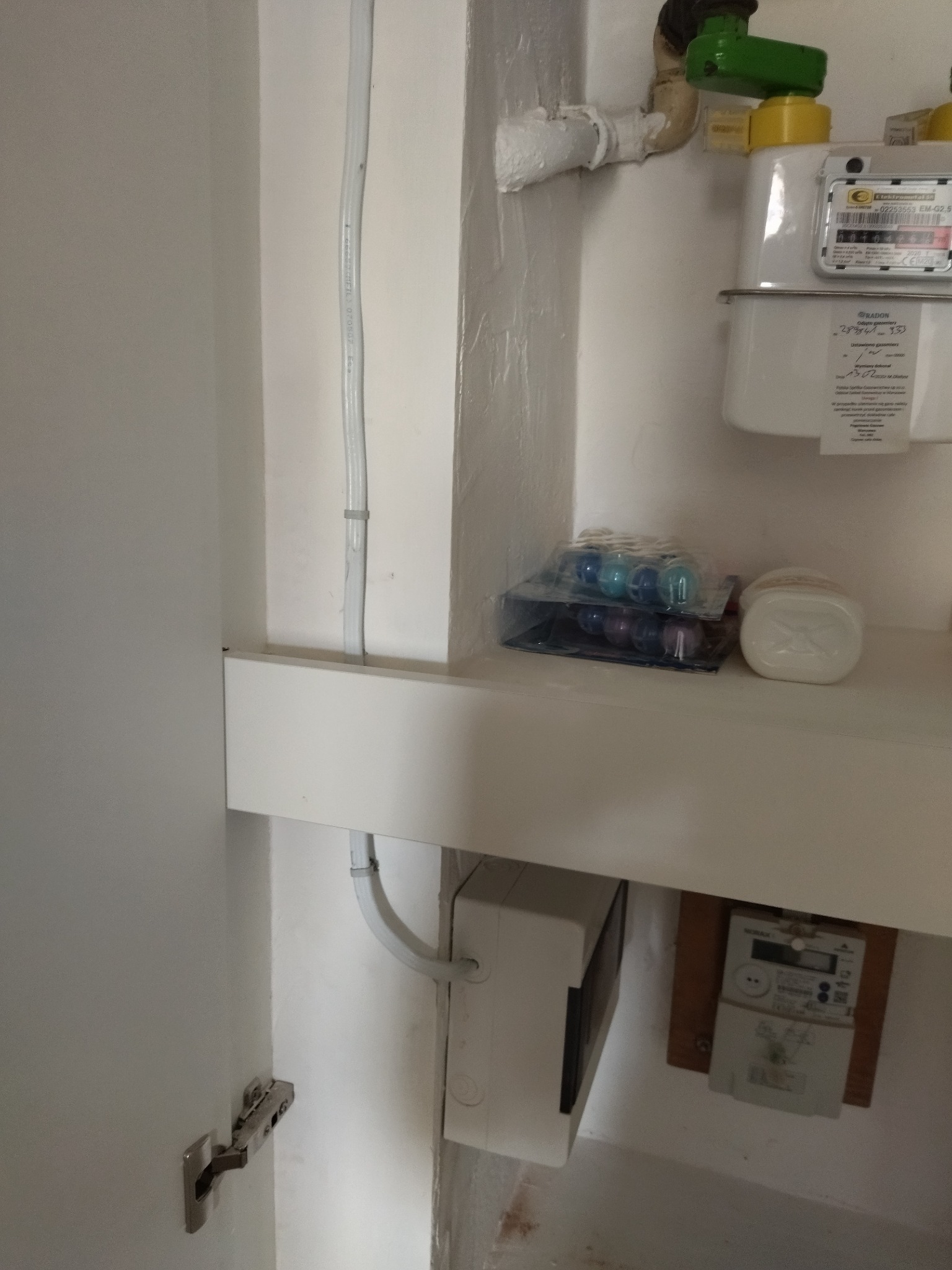 Biała instalacja elektryczna w szafce z licznikiem i rurami w Legionowie. Kabel zabezpieczony uchwytami. Widoczny licznik gazu.