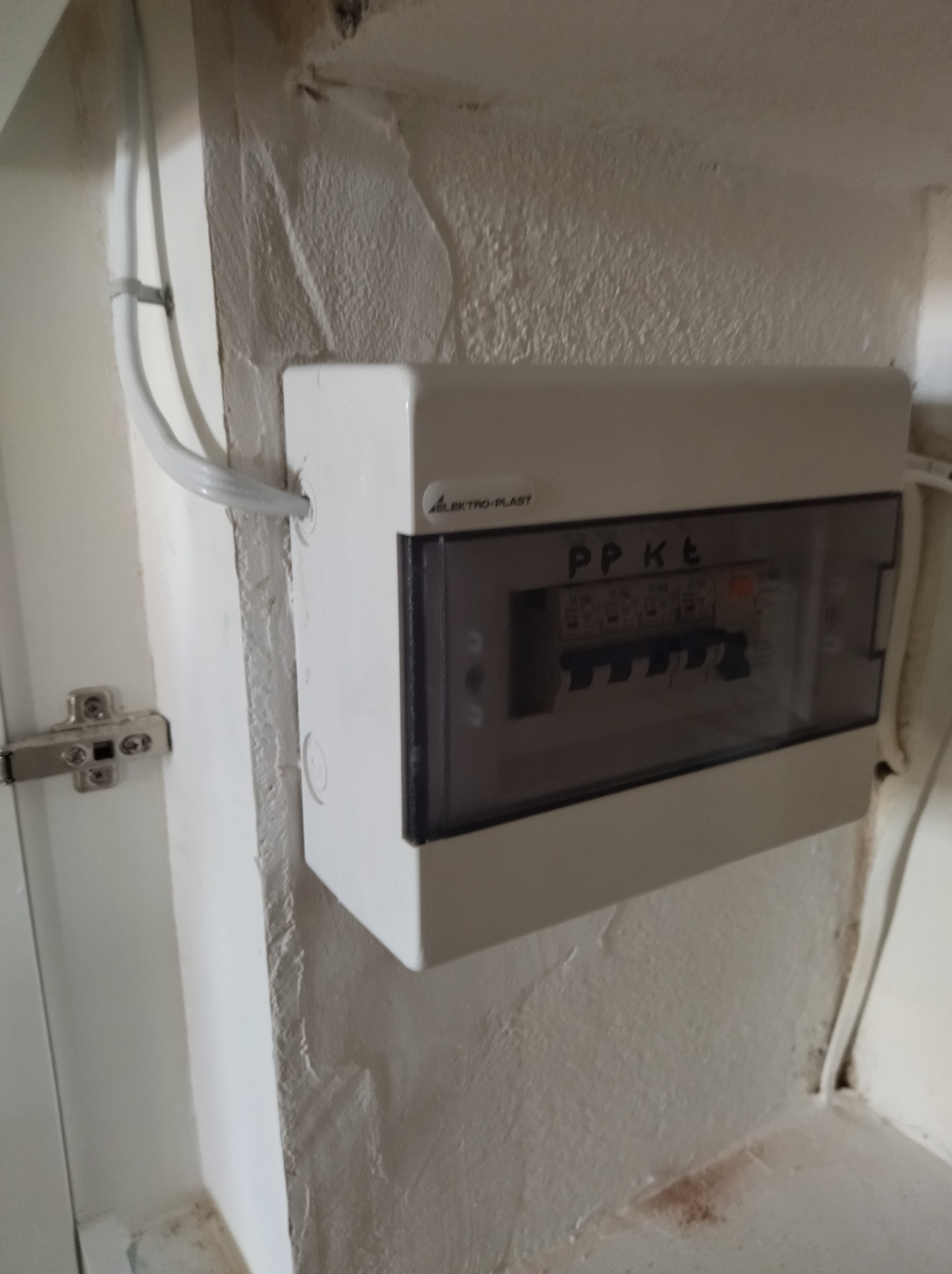 Zamknięta, biała skrzynka elektryczna PPKt firmy Elektro-Plast, zamontowana na pomalowanej ścianie. Widoczne przewody wchodzące do skrzynki i wyłączniki wewnątrz.