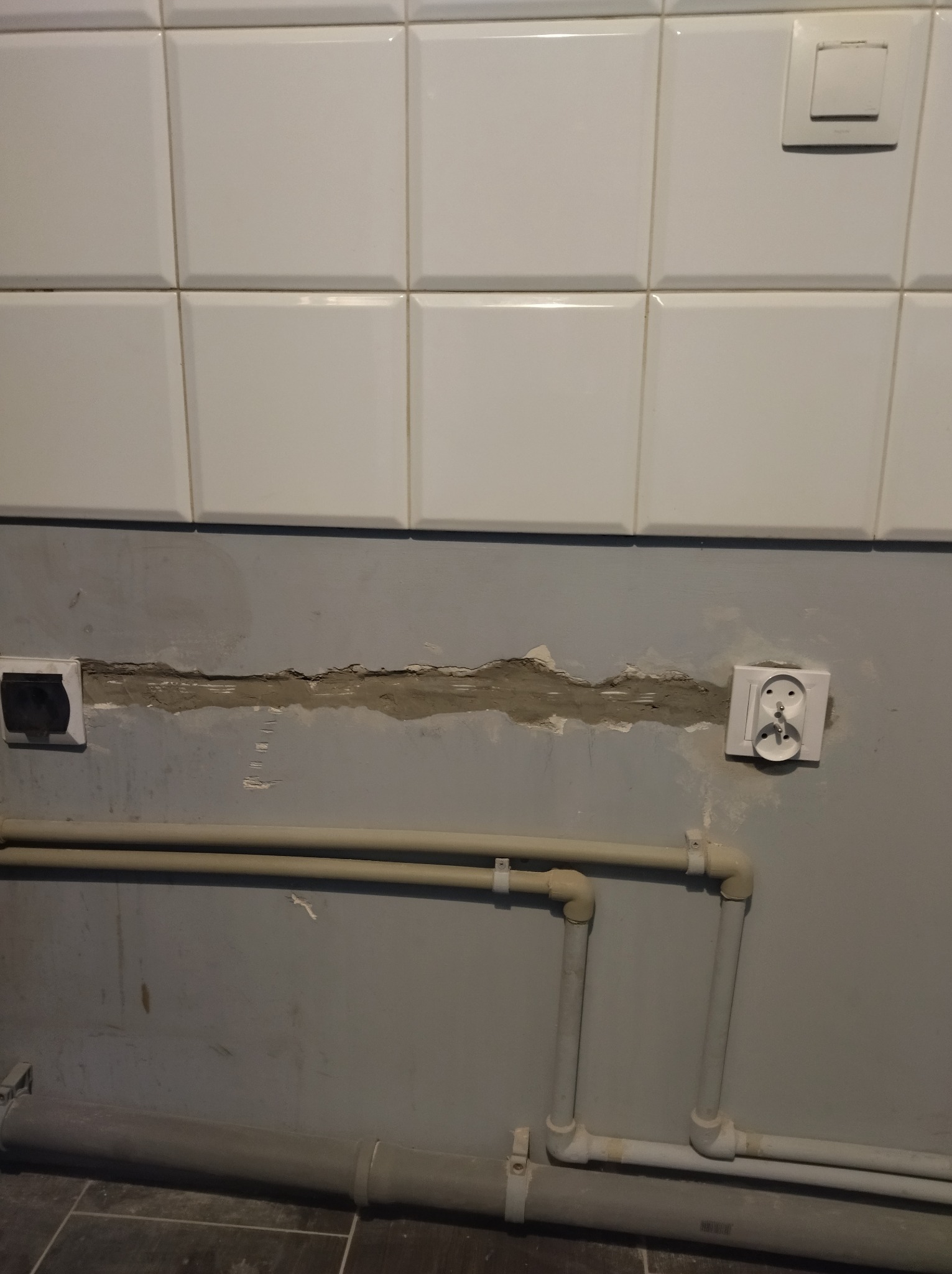 Uszkodzona ściana z gniazdkami elektrycznymi i rurami, widoczne ubytki w tynku pod płytkami. Gniazdka zamontowane po obu stronach ubytku.