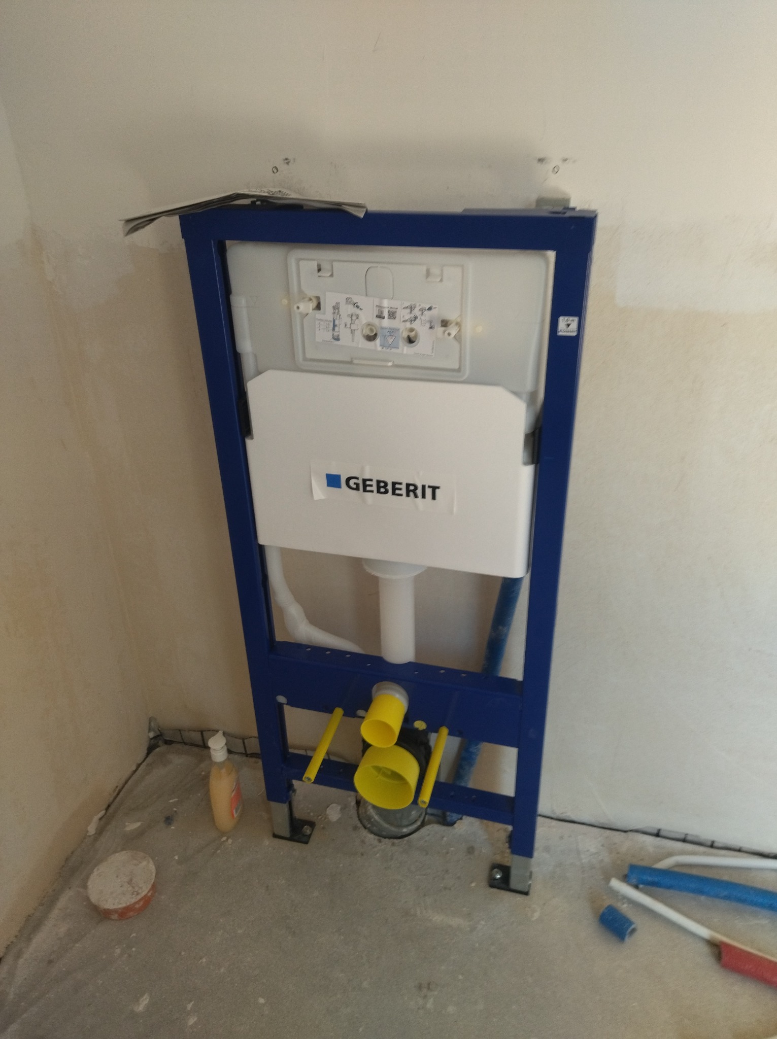 Stelaż podtynkowy Geberit w niebieskiej ramie, widoczne białe rury i żółte elementy instalacji. W tle surowa ściana i podłoga w trakcie prac wykończeniowych.