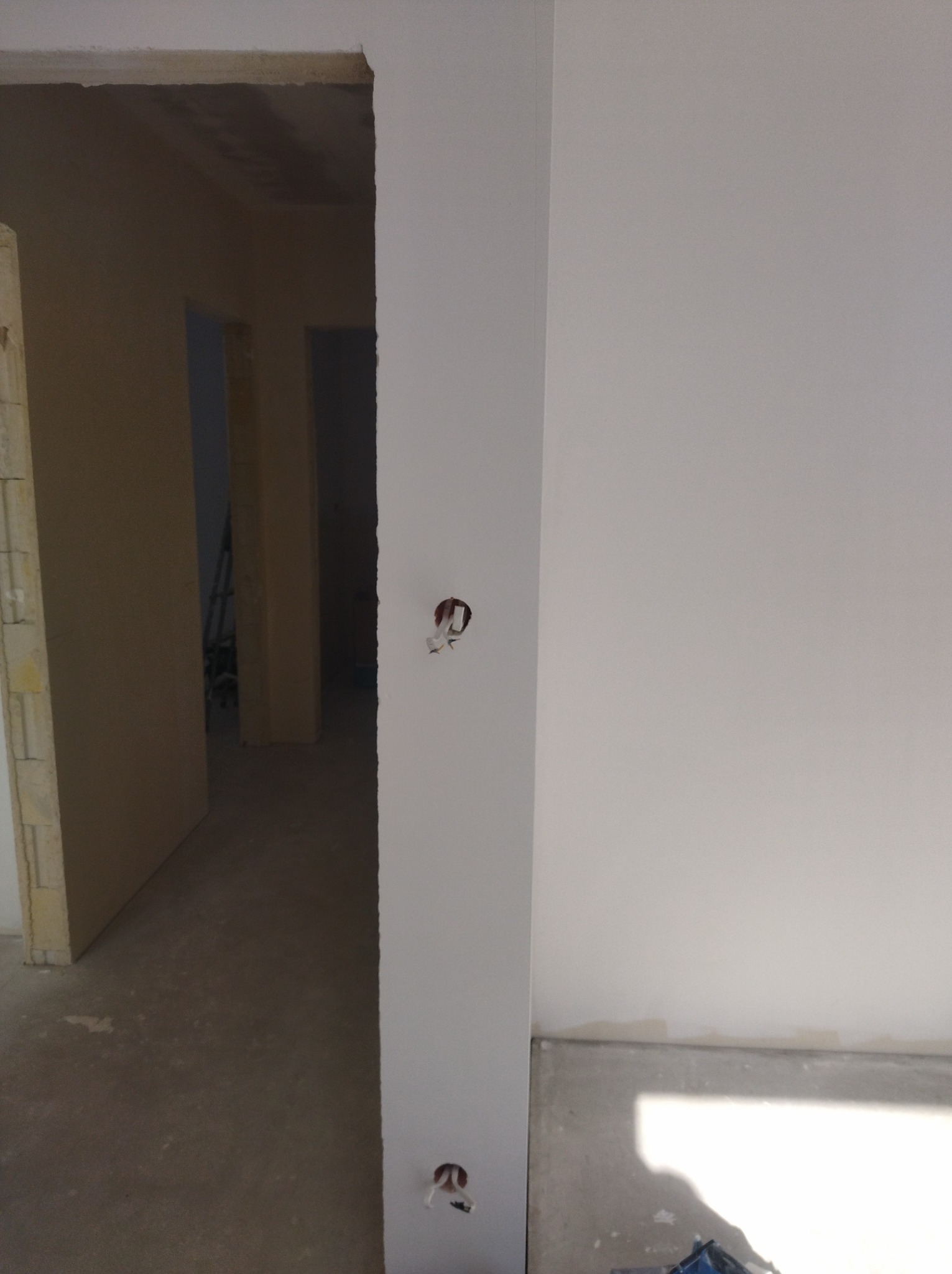 Biała ściana z dwoma otworami na gniazdka elektryczne, widok na surowy korytarz w tle. Świeżo pomalowane ściany, brak osprzętu.
