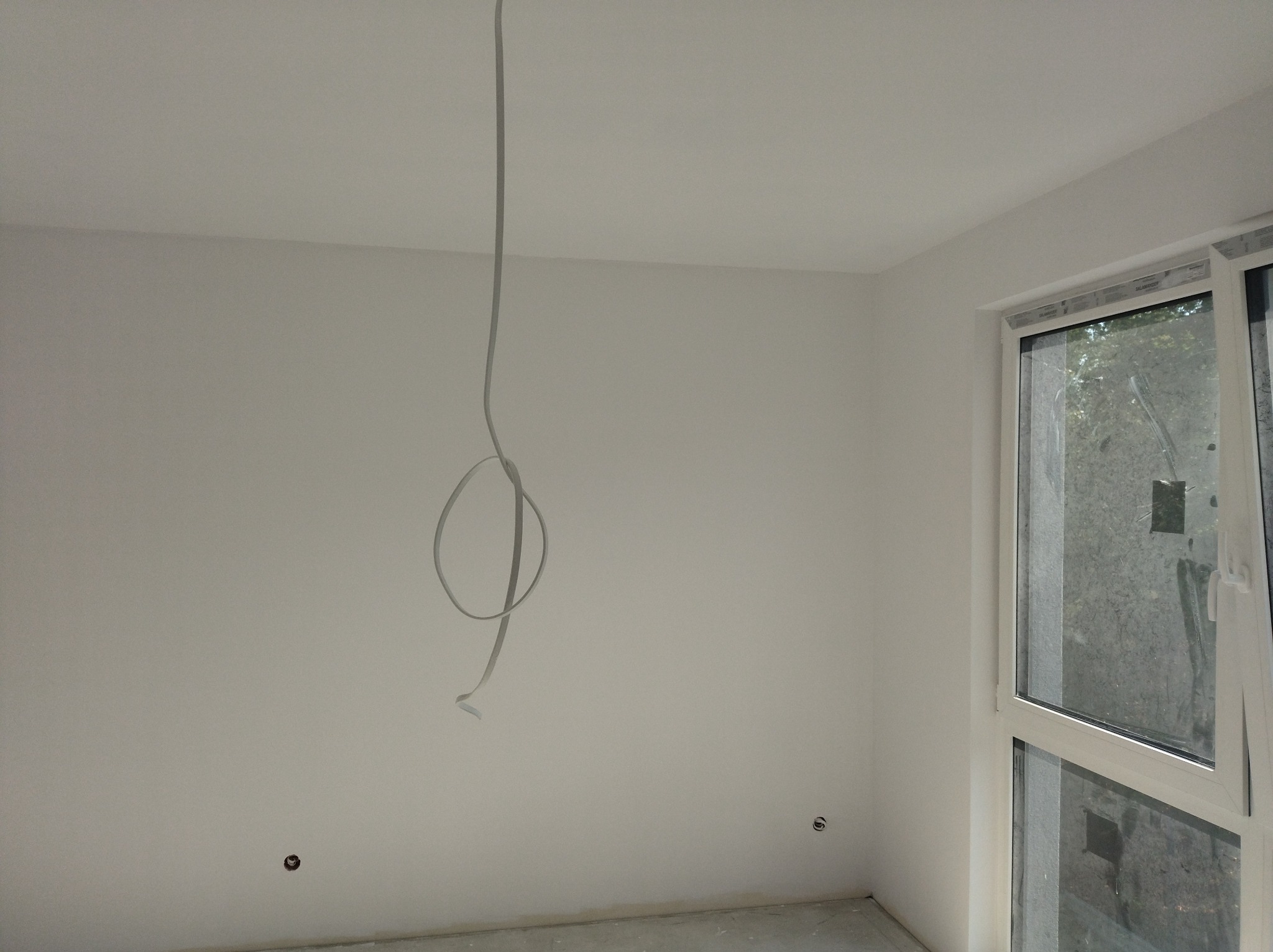 Szary kabel zwisa z sufitu w jasnym pokoju z oknem. Widoczne puszki elektryczne na ścianie. Przygotowanie do instalacji oświetlenia w nowym mieszkaniu.